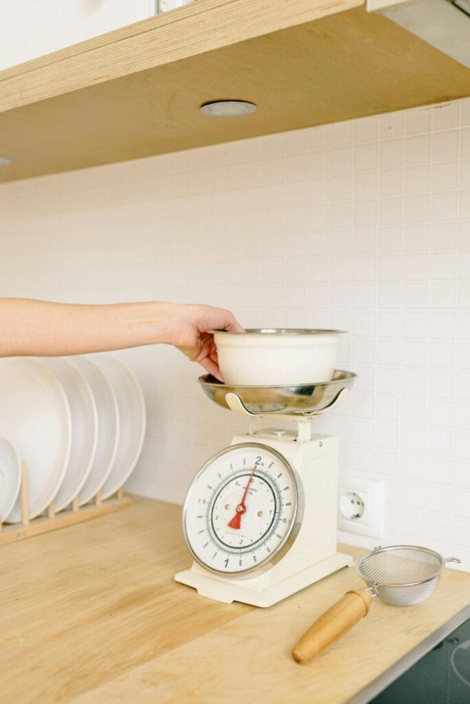 A hand placing a bowl on a retro kitchen scale in a modern kitchen setting.